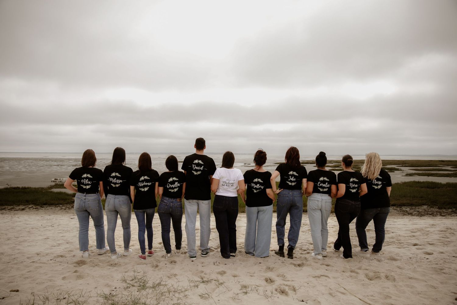 A group stands with their backs to camera on a beach wearing matching black and white shirts on an overcast day.