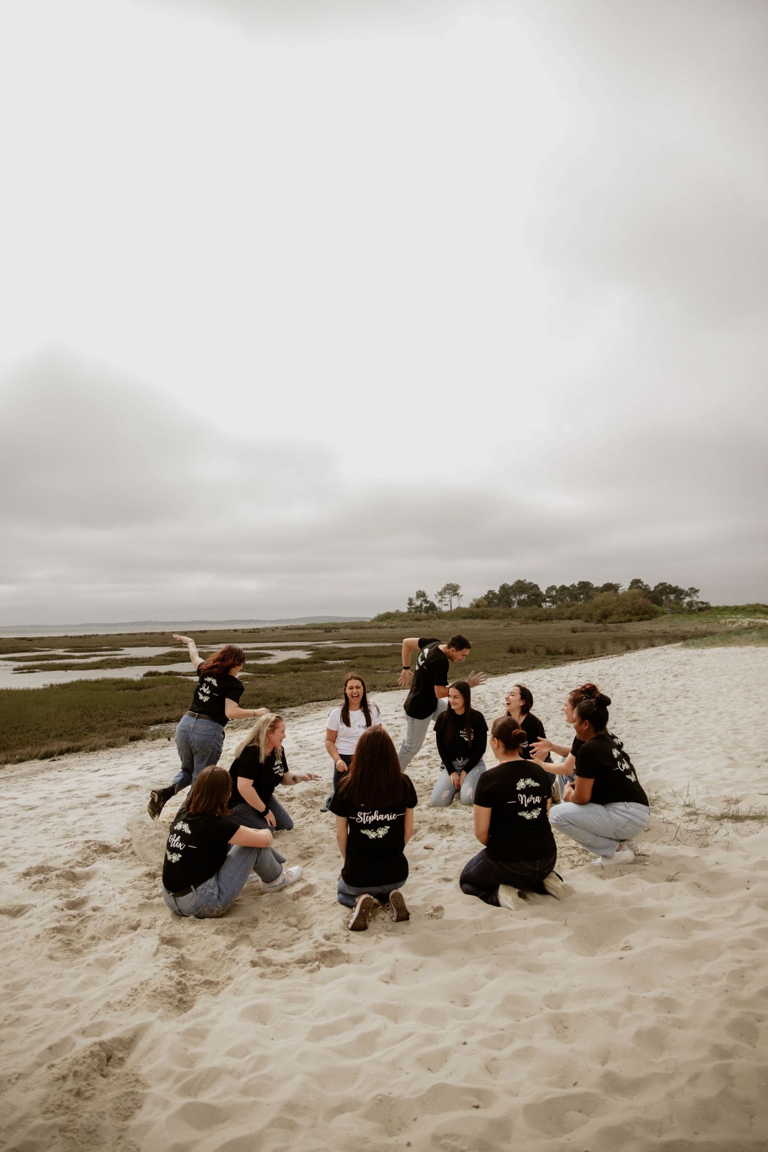 Group of people wearing matching black shirts gather on a sandy beach under an overcast sky.