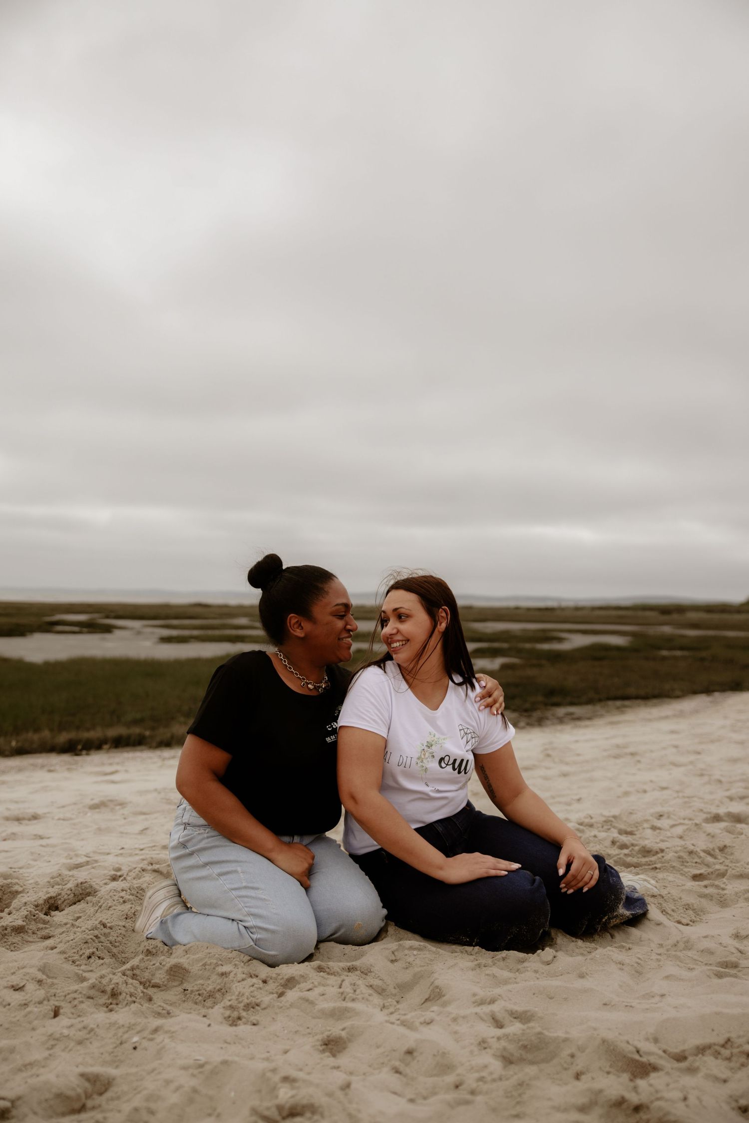 Two people sit together laughing and sharing a joyful moment on a sandy beach under an overcast sky.