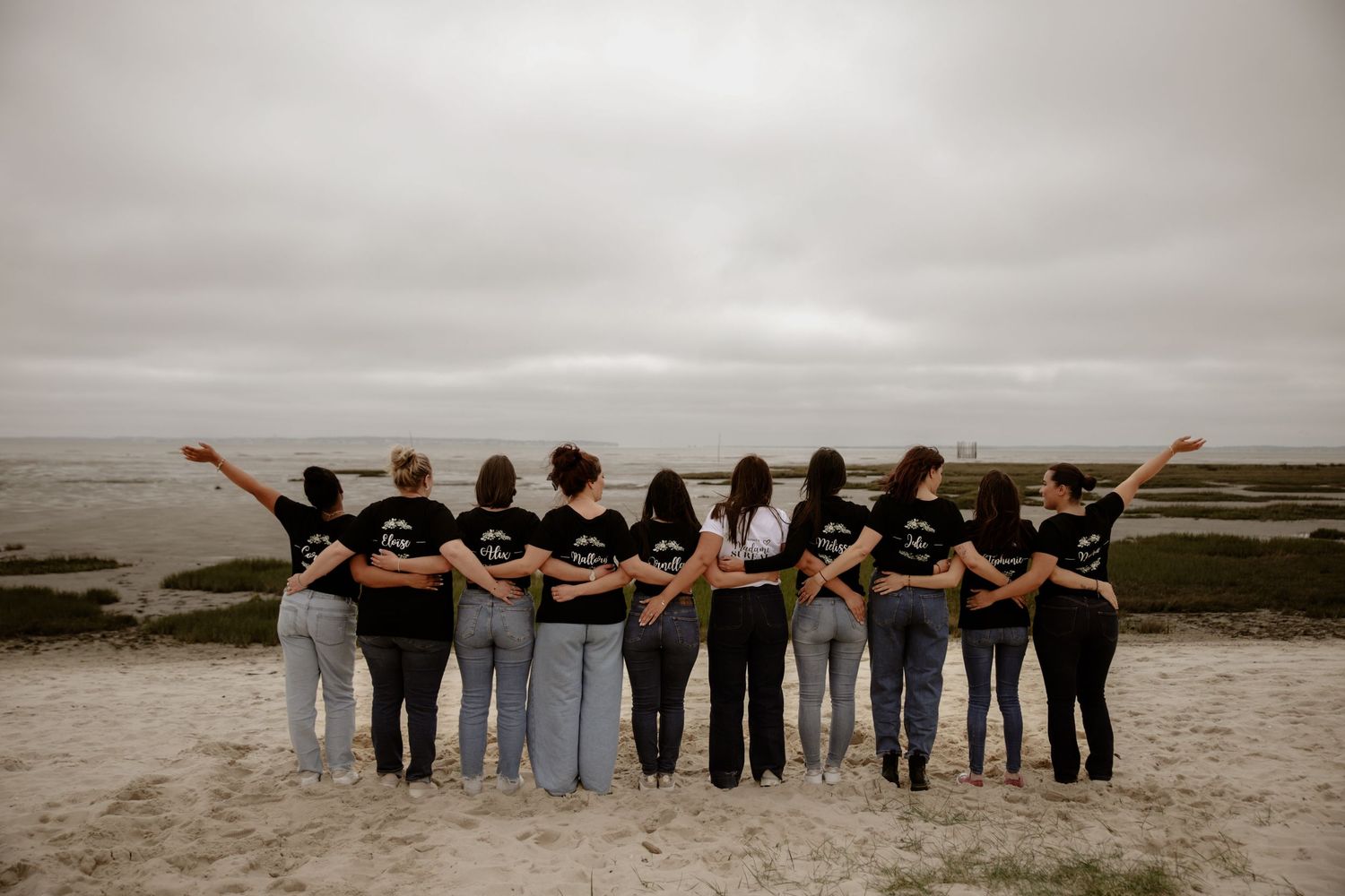 A group in matching black shirts stands with their backs to the camera while overlooking a moody beach landscape.
