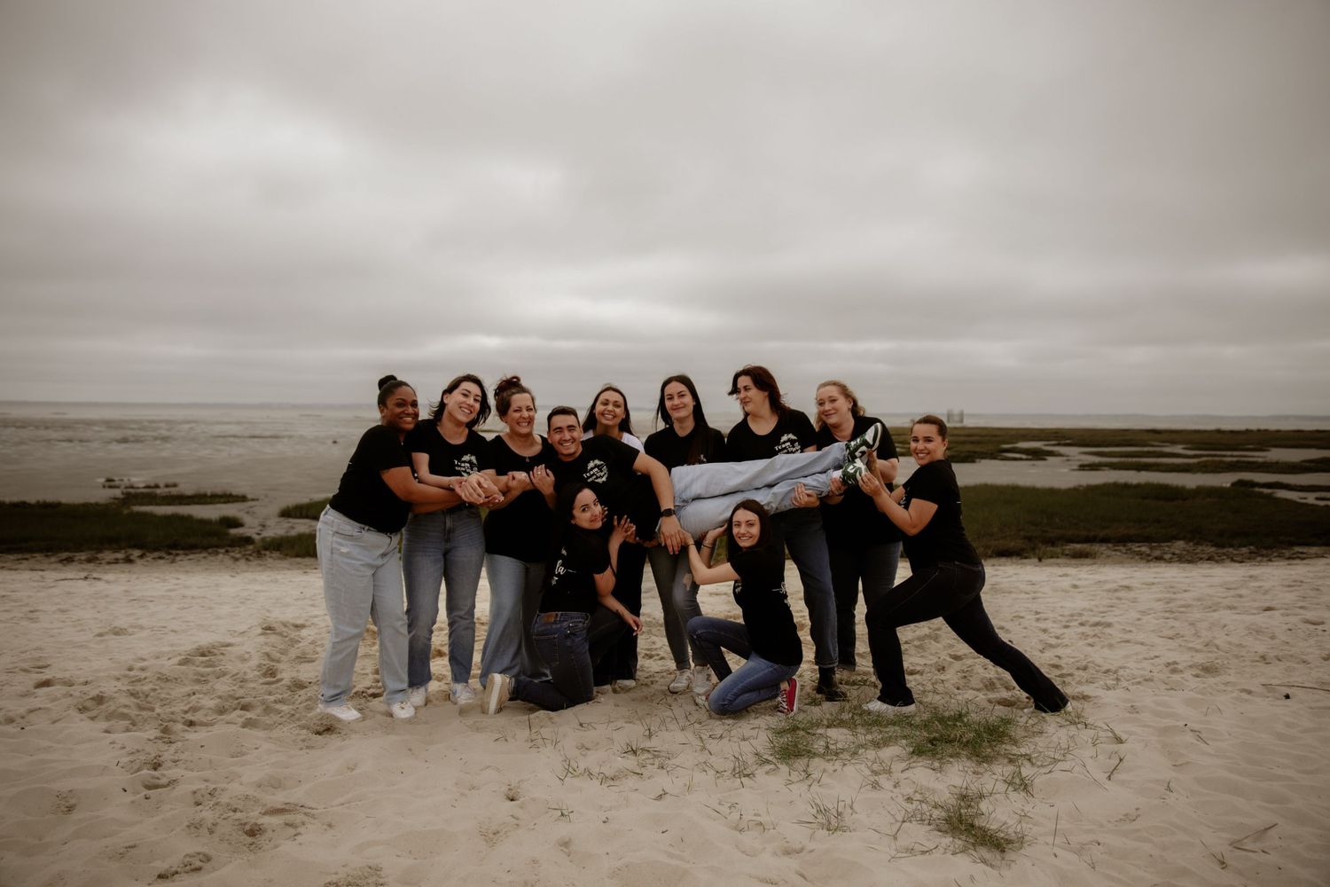 A group poses together on a cloudy beach with dramatic overcast skies and sand dunes in the background.