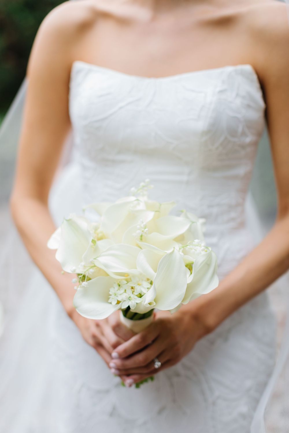 A stunning white calla lily wedding bouquet held against a white strapless wedding gown in soft natural lighting.