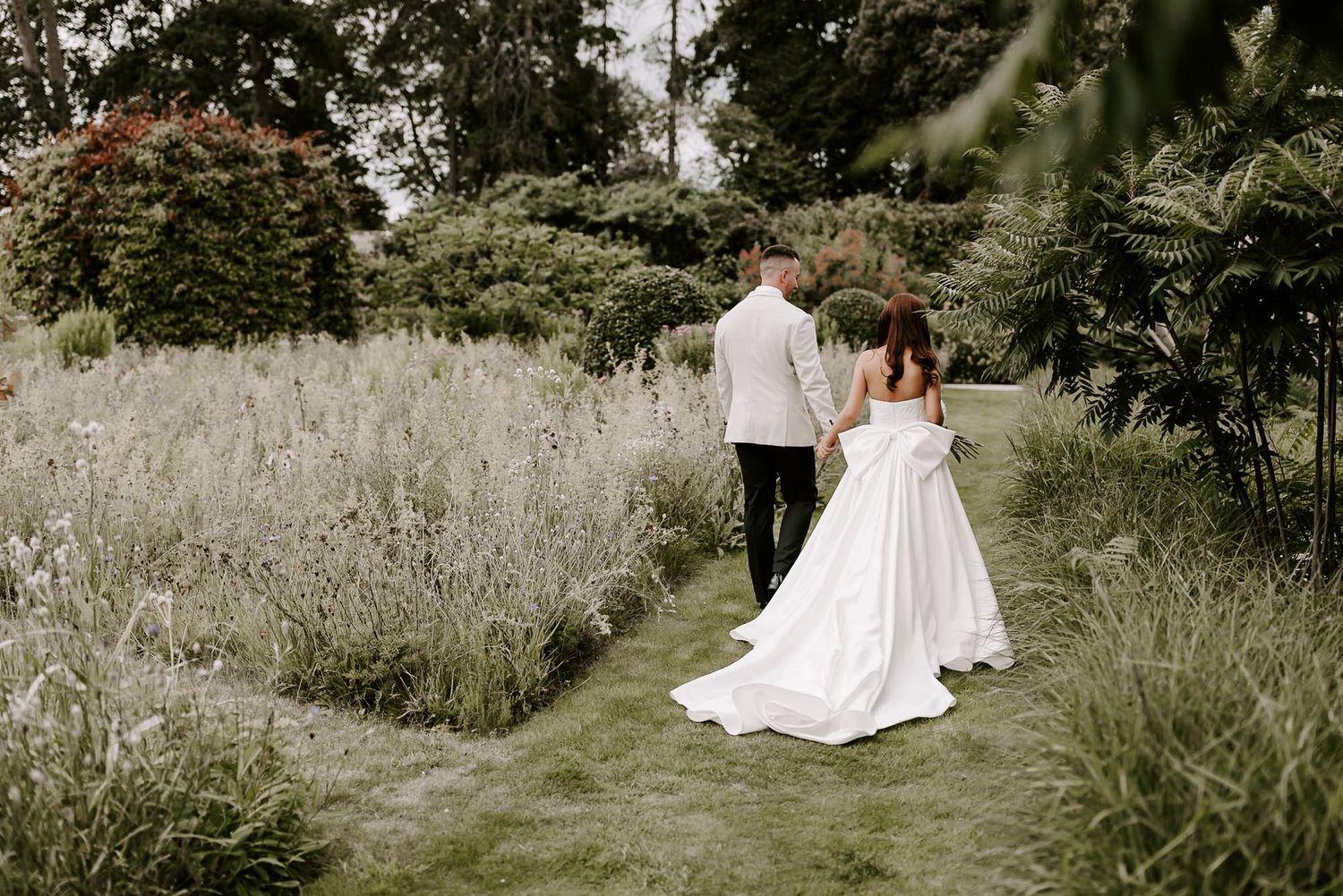 A couple in wedding attire walk hand in hand through a rustic field of tall grass on their special day.