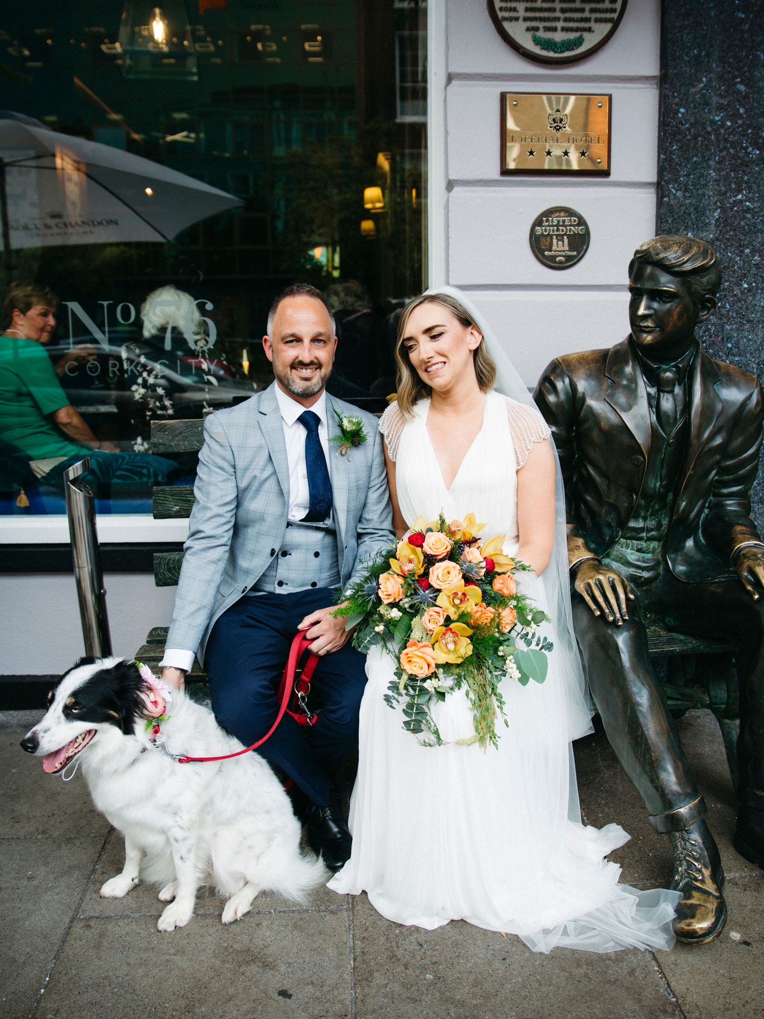 Newlyweds pose with their white dog next to a bronze statue outside a storefront on their wedding day.