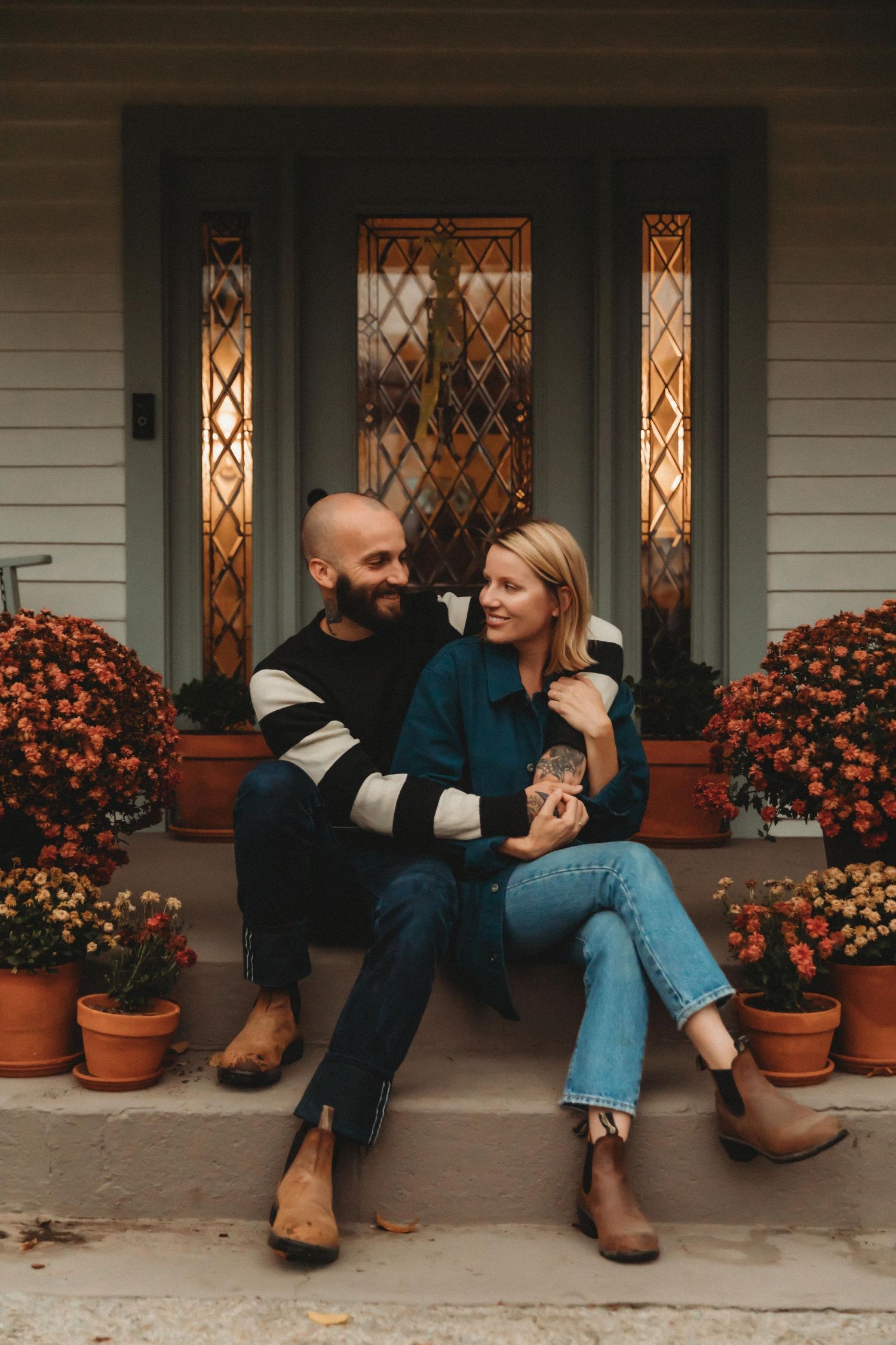 Couple sitting on front porch steps surrounded by orange mums in autumn with decorative door behind them.