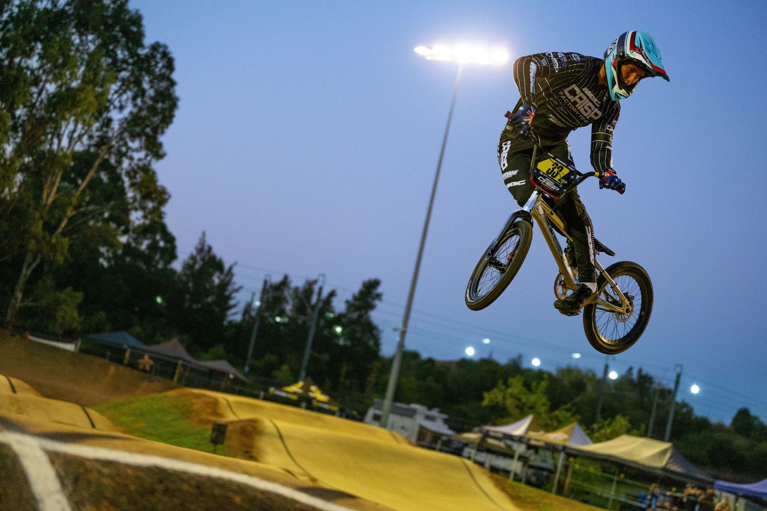 BMX rider performs aerial jump silhouetted against evening sky at illuminated dirt track course.