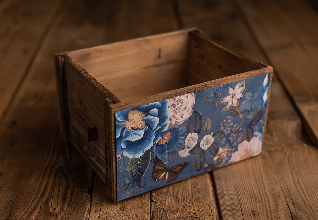 A rustic wooden crate adorned with blue floral and butterfly fabric trim sitting on a dark hardwood floor.