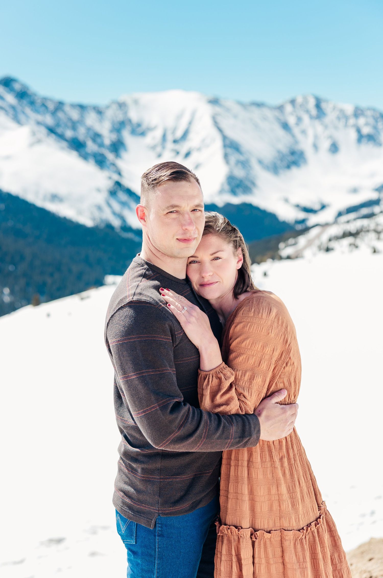 A couple embraces while standing in snow with majestic snow-capped Rocky Mountains rising behind them on a bright winter day.