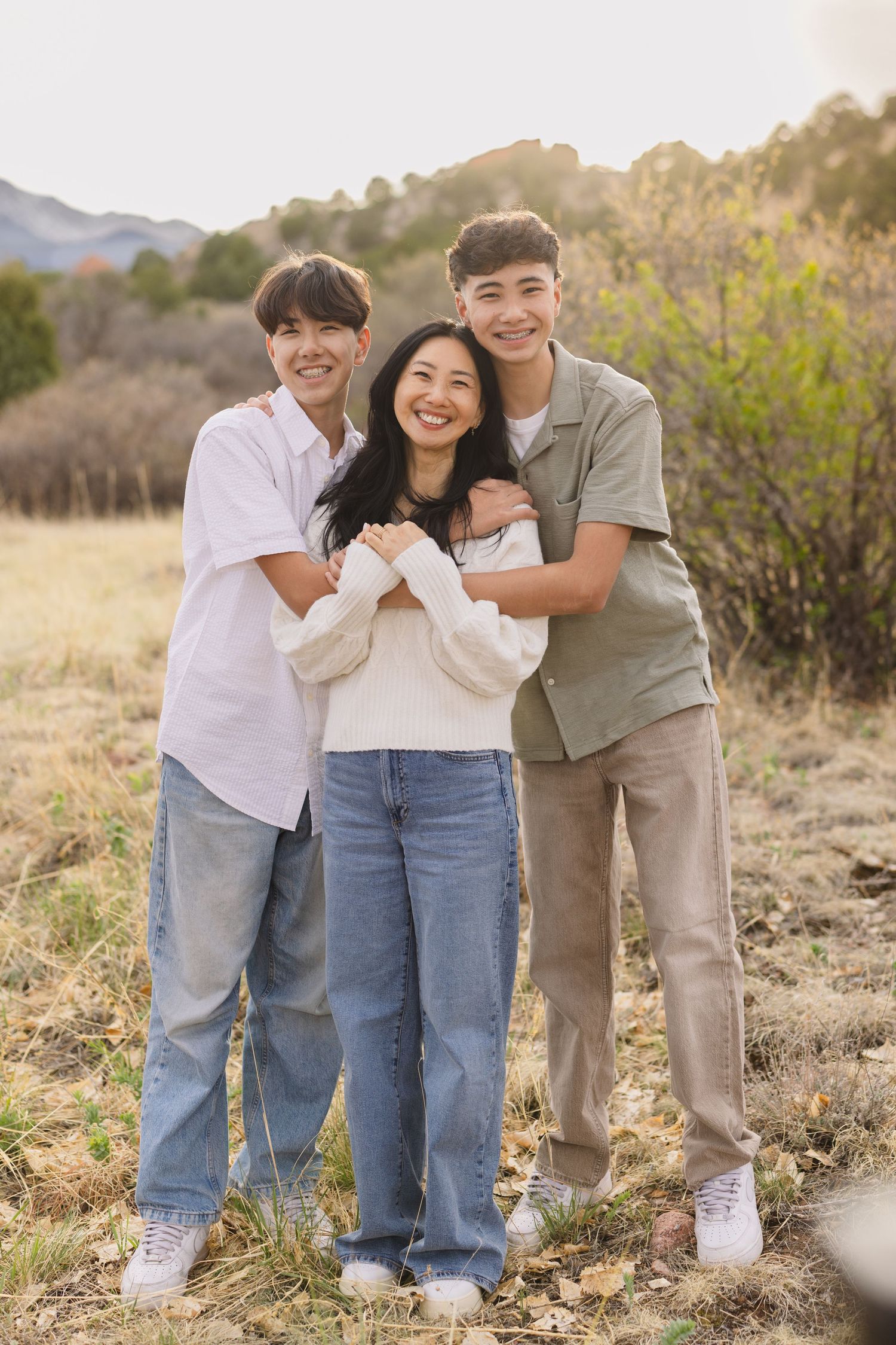 A smiling mother stands between her two teenage sons hugging her in a sunny outdoor field with mountains behind.