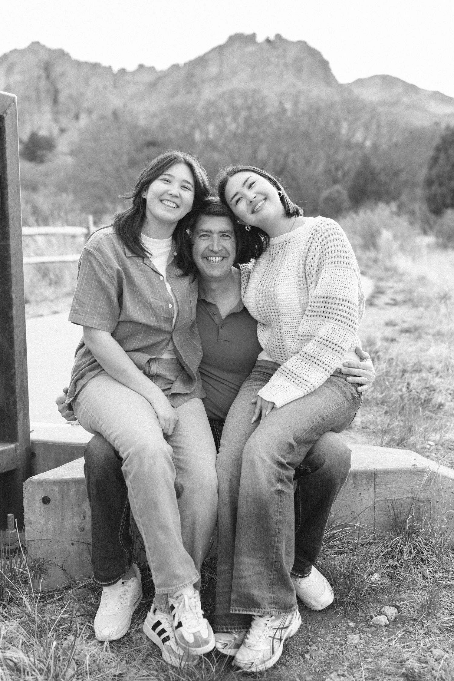 A father sits between two daughters leaning on him on a bench with rocky mountains in this black and white portrait.