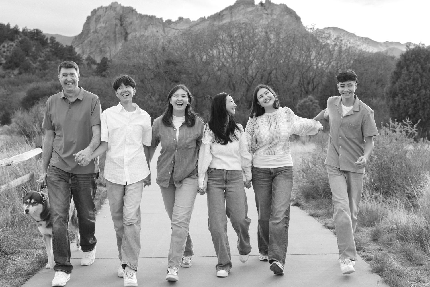 Black and white photo of a joyful family of six walking together on a path with rocky mountains in the background.