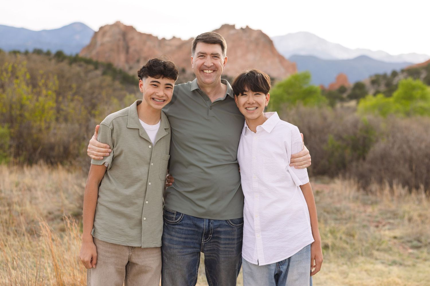 A smiling man and two teenage boys pose together outdoors with rocky mountain formations and greenery in the background.