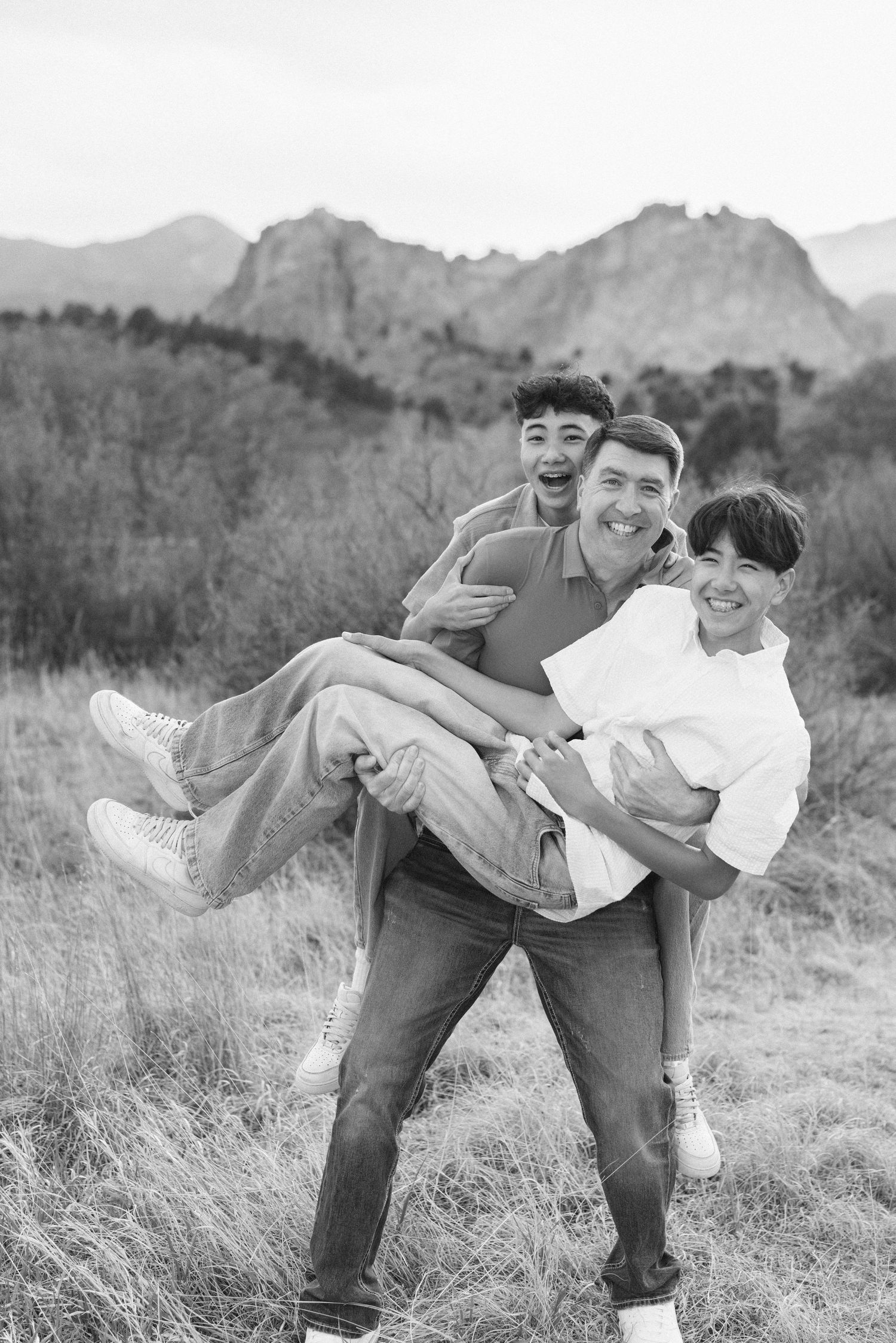 A man joyfully carries a woman and teen boy in a field with mountain views in this black and white family photo.