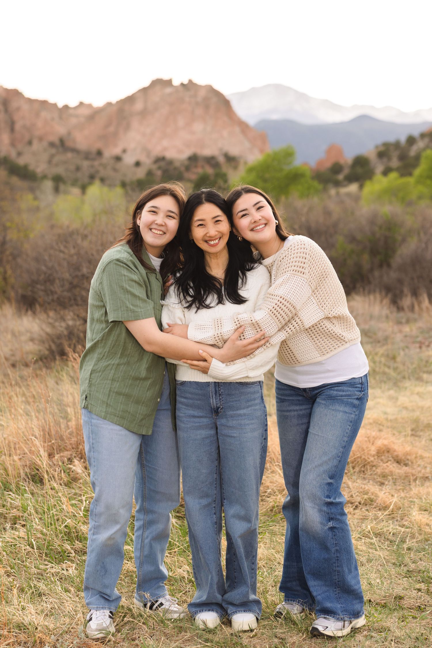 Three young women smiling and embracing outdoors with rocky mountain landscape in background.