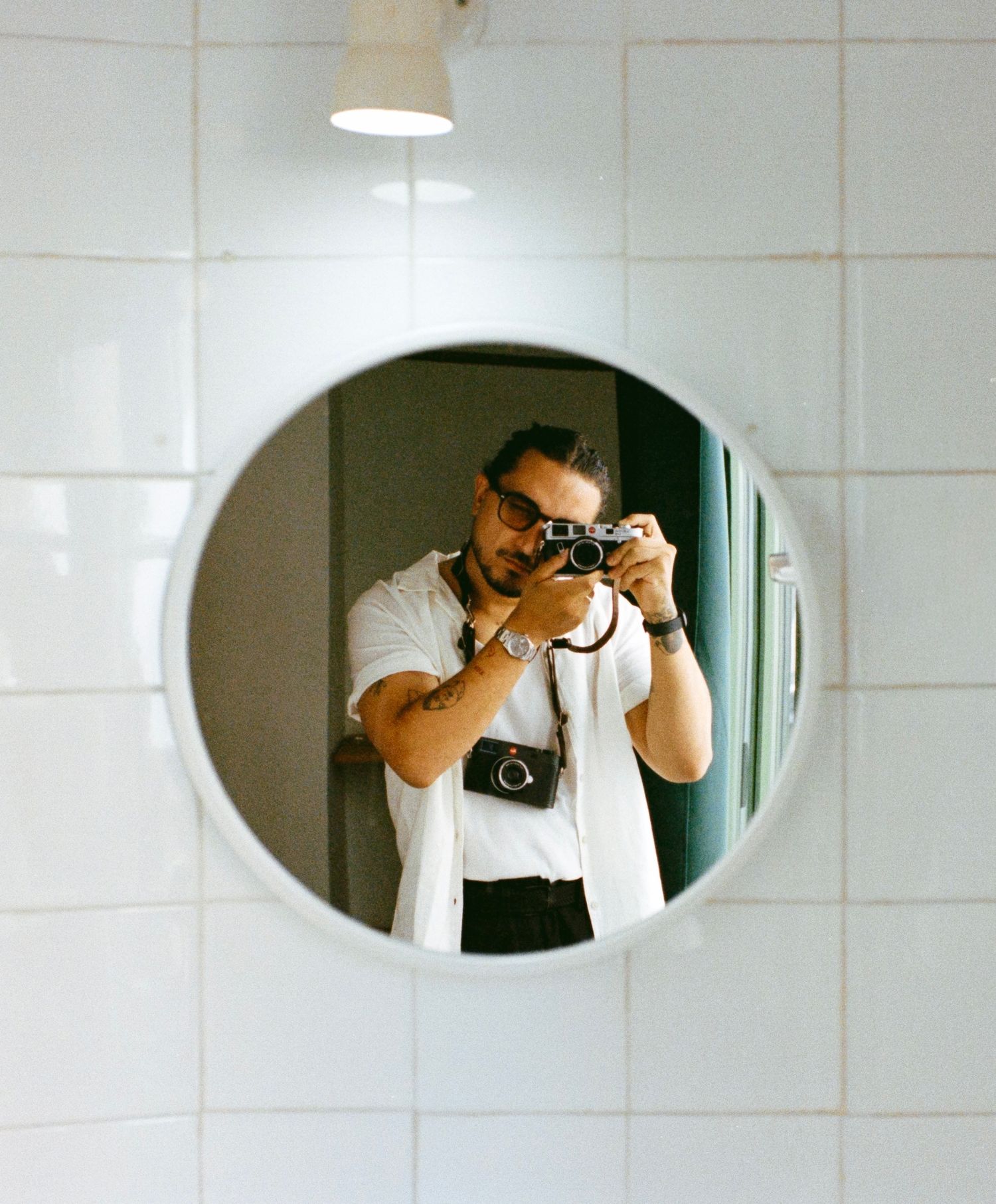 Man taking mirror selfie with film camera in round bathroom mirror, wearing white shirt and sunglasses.