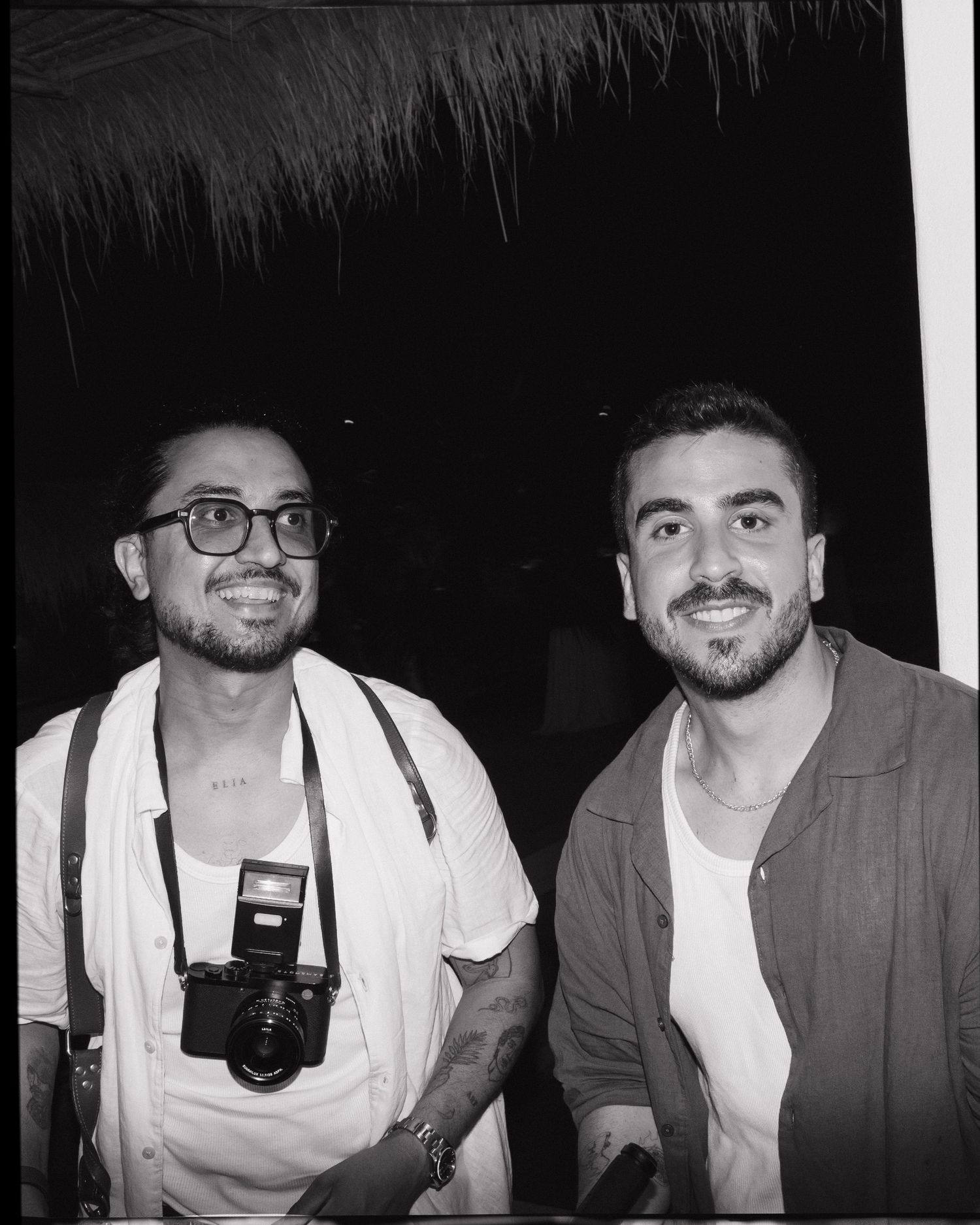 Two smiling men pose together outdoors at night, one wearing glasses with a camera around his neck.