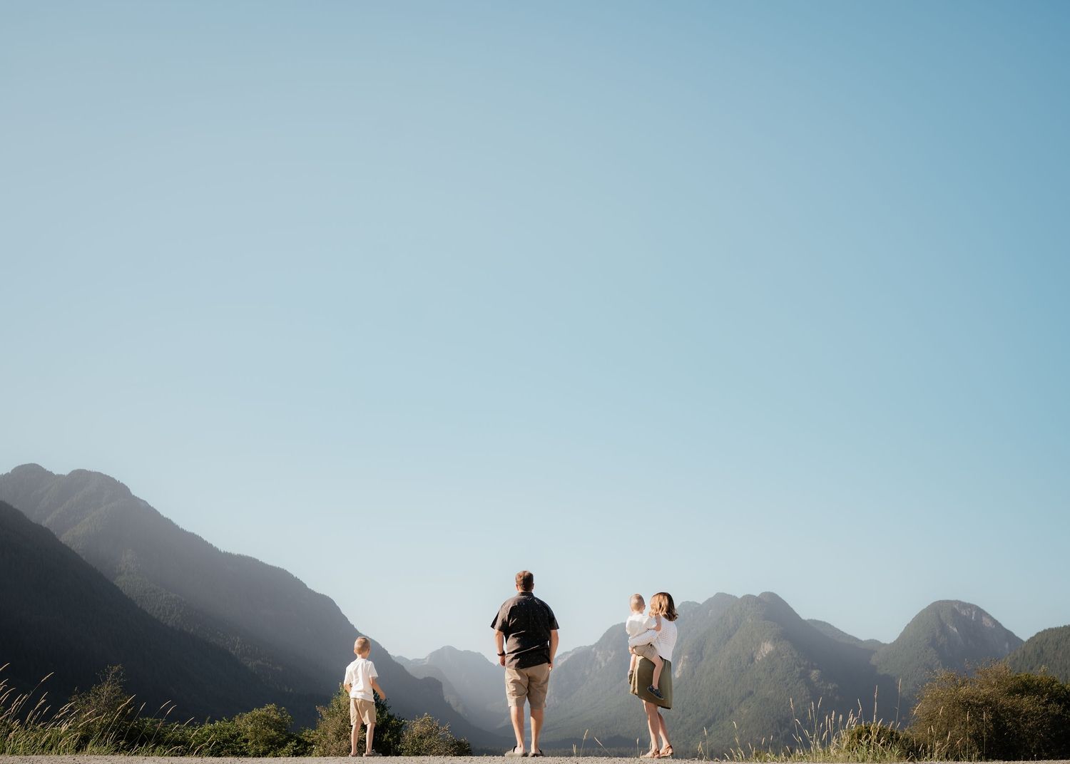 People and a dog walk along a mountain trail with dramatic mountain peaks silhouetted against a blue sky.