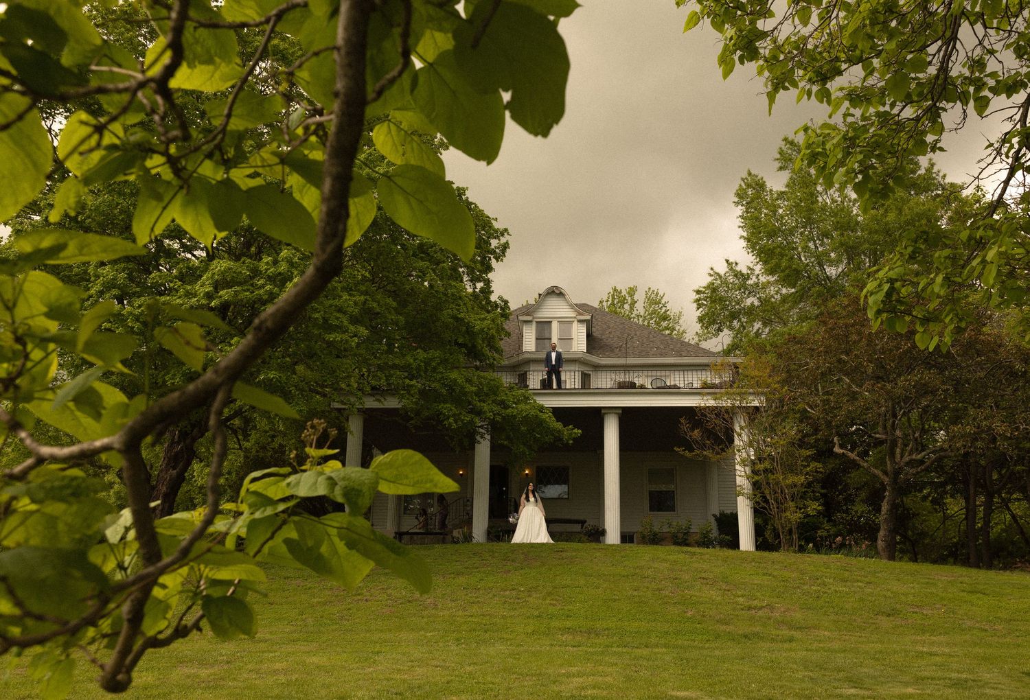 A white colonial house with columns sits on a green lawn surrounded by lush trees on an overcast day.