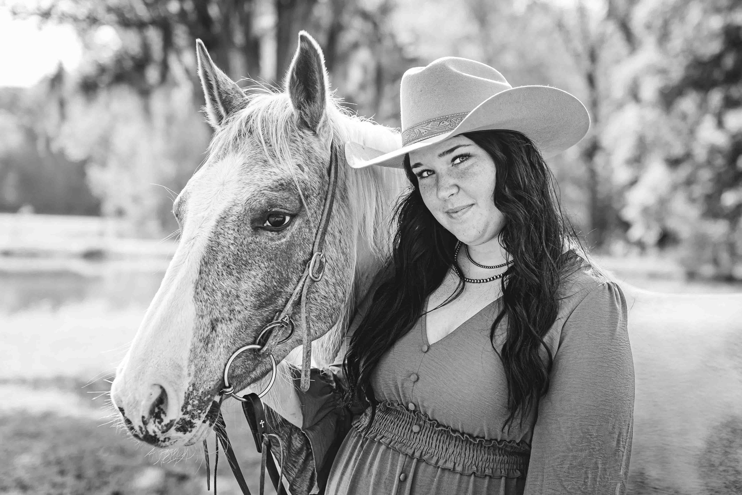A western-style black and white portrait featuring a woman in a cowboy hat standing beside a white horse in nature.