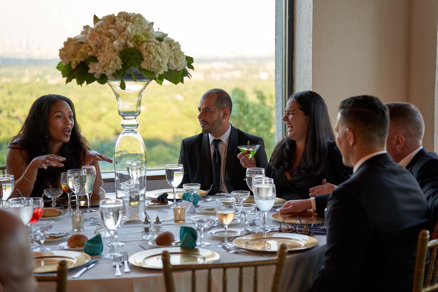 Guests in formal attire conversing at elegantly set dining table with city view and floral centerpiece.