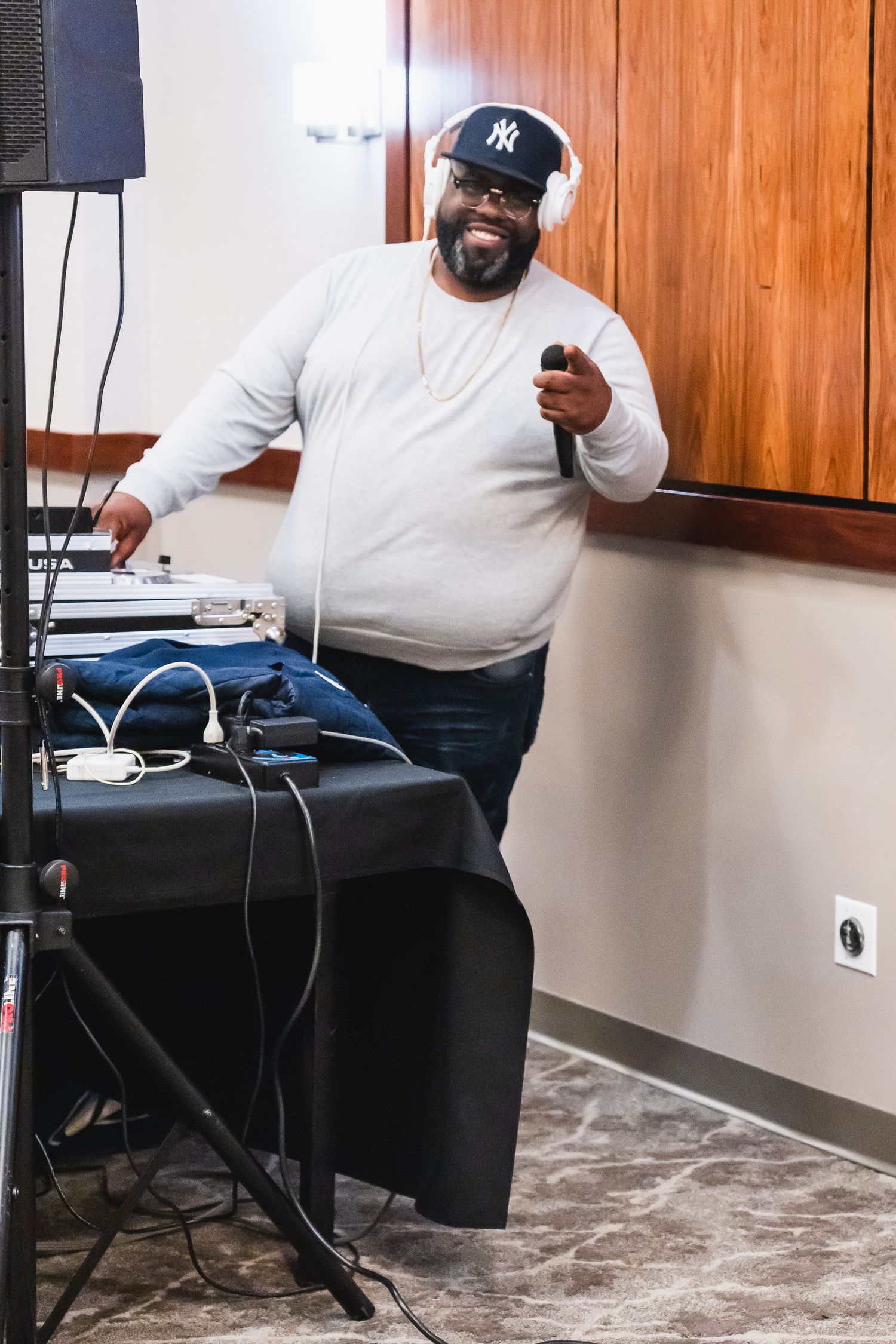 A smiling DJ wearing headphones and NY cap operates equipment at an indoor event venue.