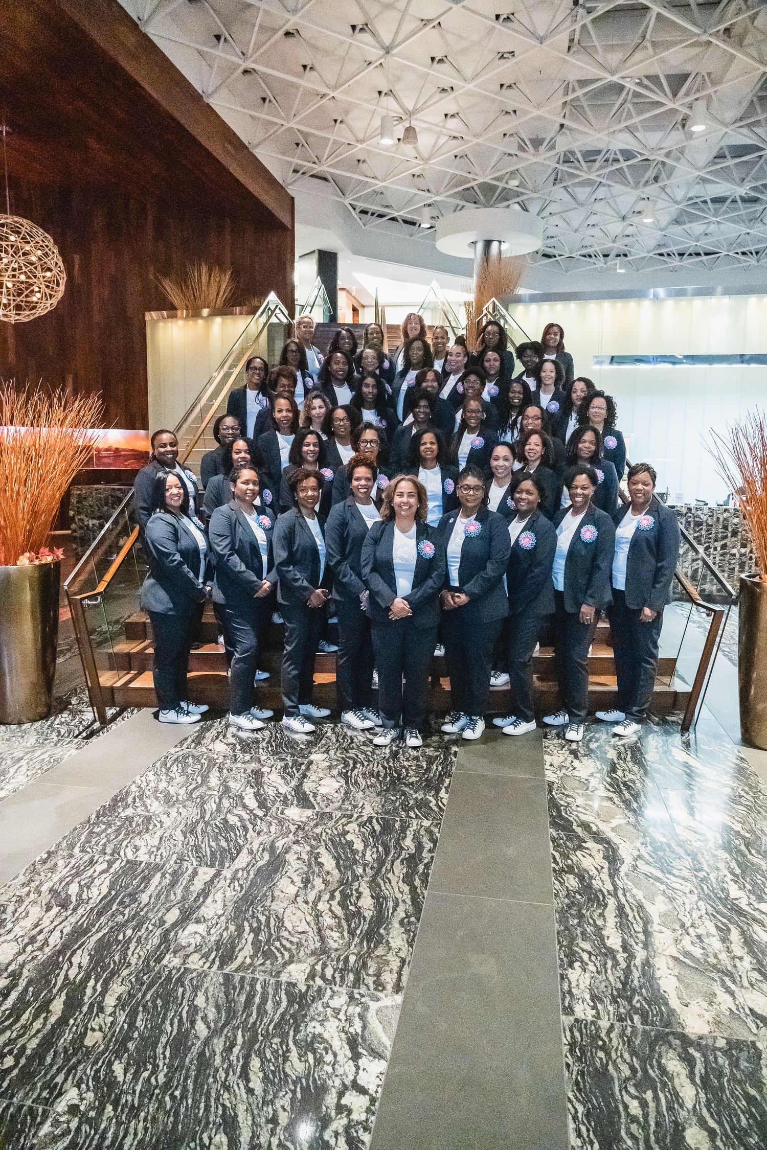 Large group of professional women in matching navy blazers posing on grand staircase in modern lobby.