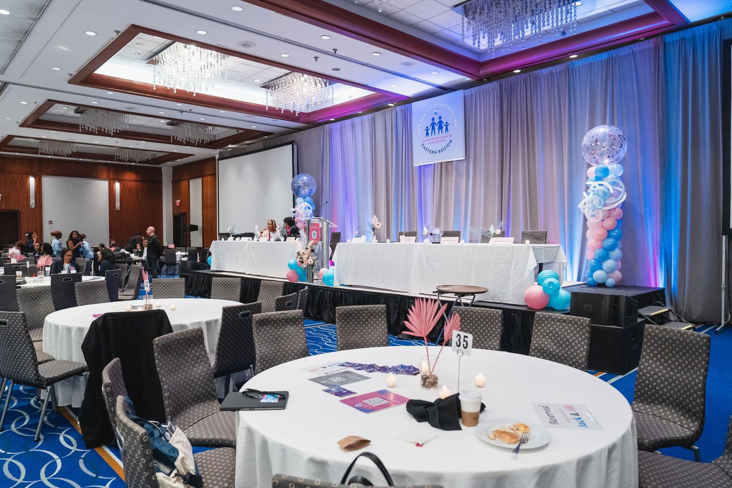 An elegantly decorated conference ballroom with round tables, balloon centerpieces and blue lighting.