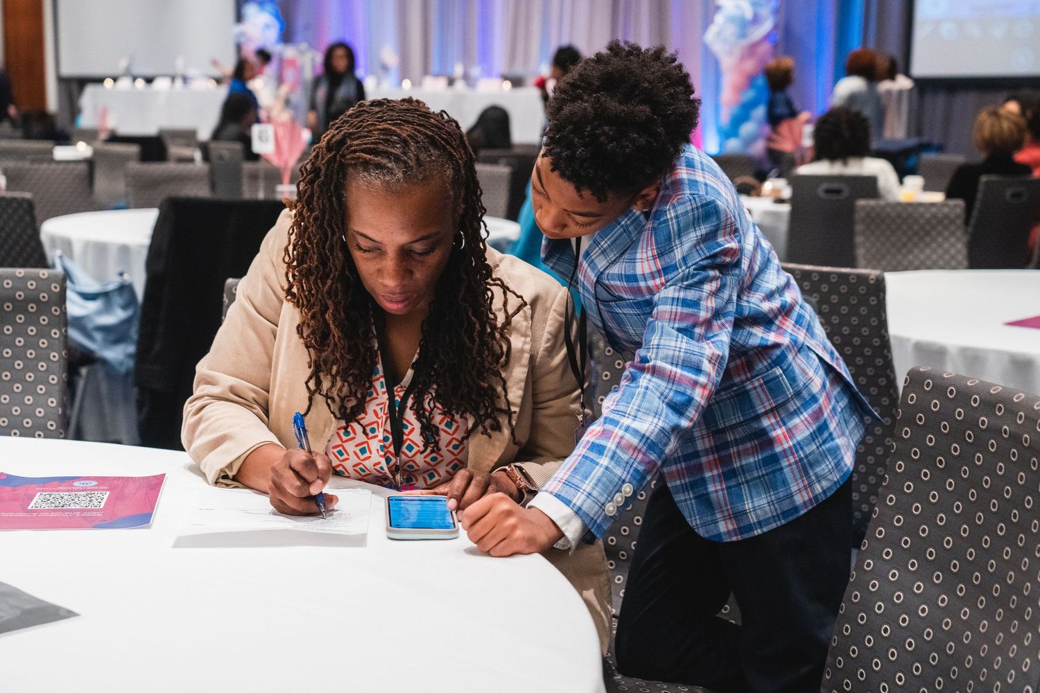 Two women collaborate at a round table, writing and reviewing documents at a professional conference.