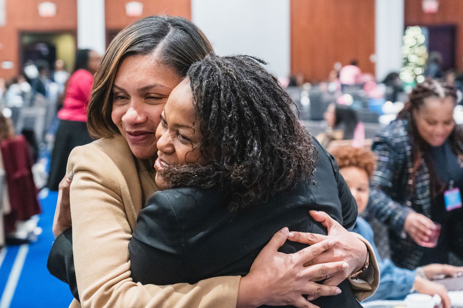 Two women share a warm embrace at an indoor community gathering event.