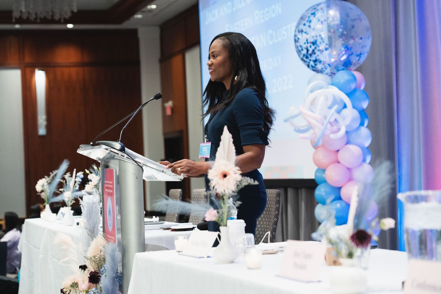 Professional woman speaking at podium during formal event with balloon decorations in background.