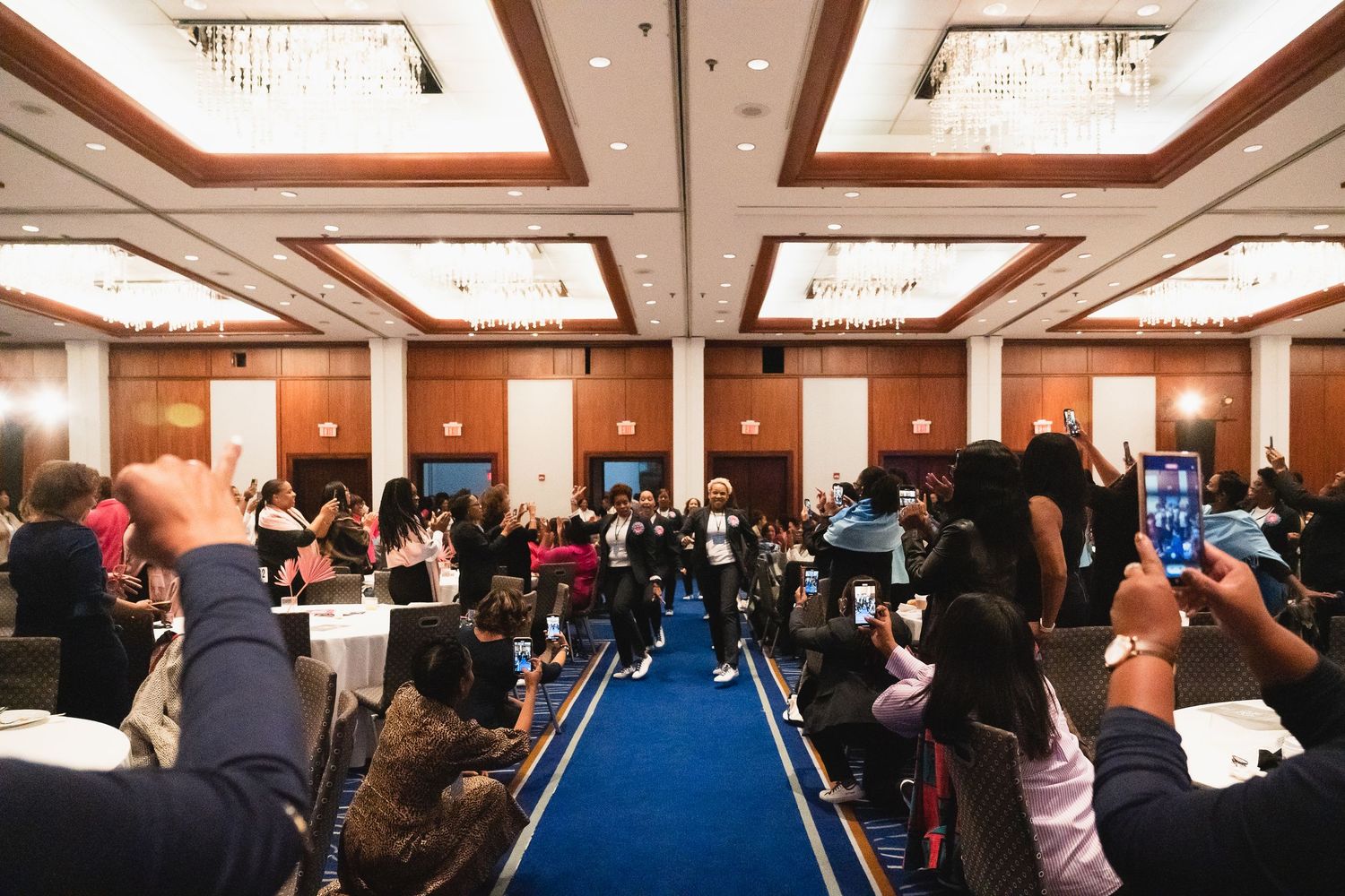 A large crowd gathers in a grand ballroom for a professional conference event with blue carpet runway.