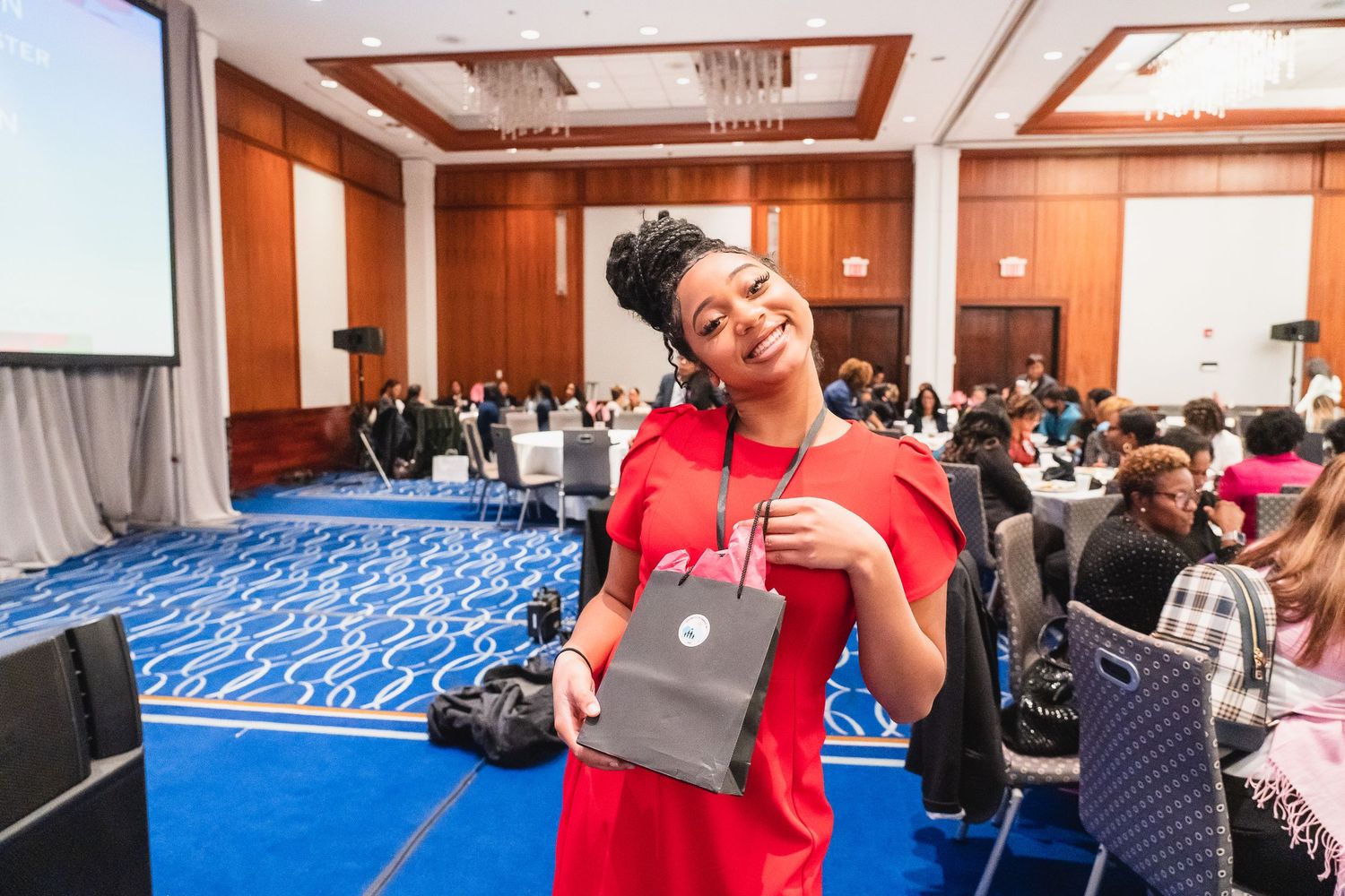 A woman in a red dress smiles while holding a gray gift bag at a professional conference event.