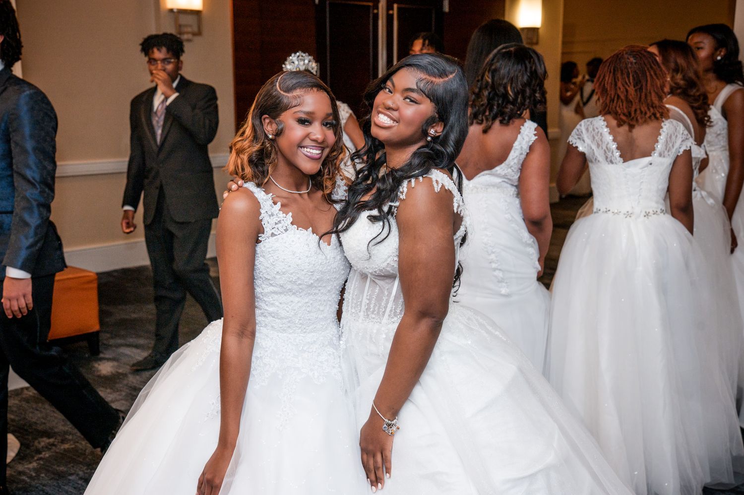 Two young Black women in white ball gowns and tiaras smiling together at a debutante ball.