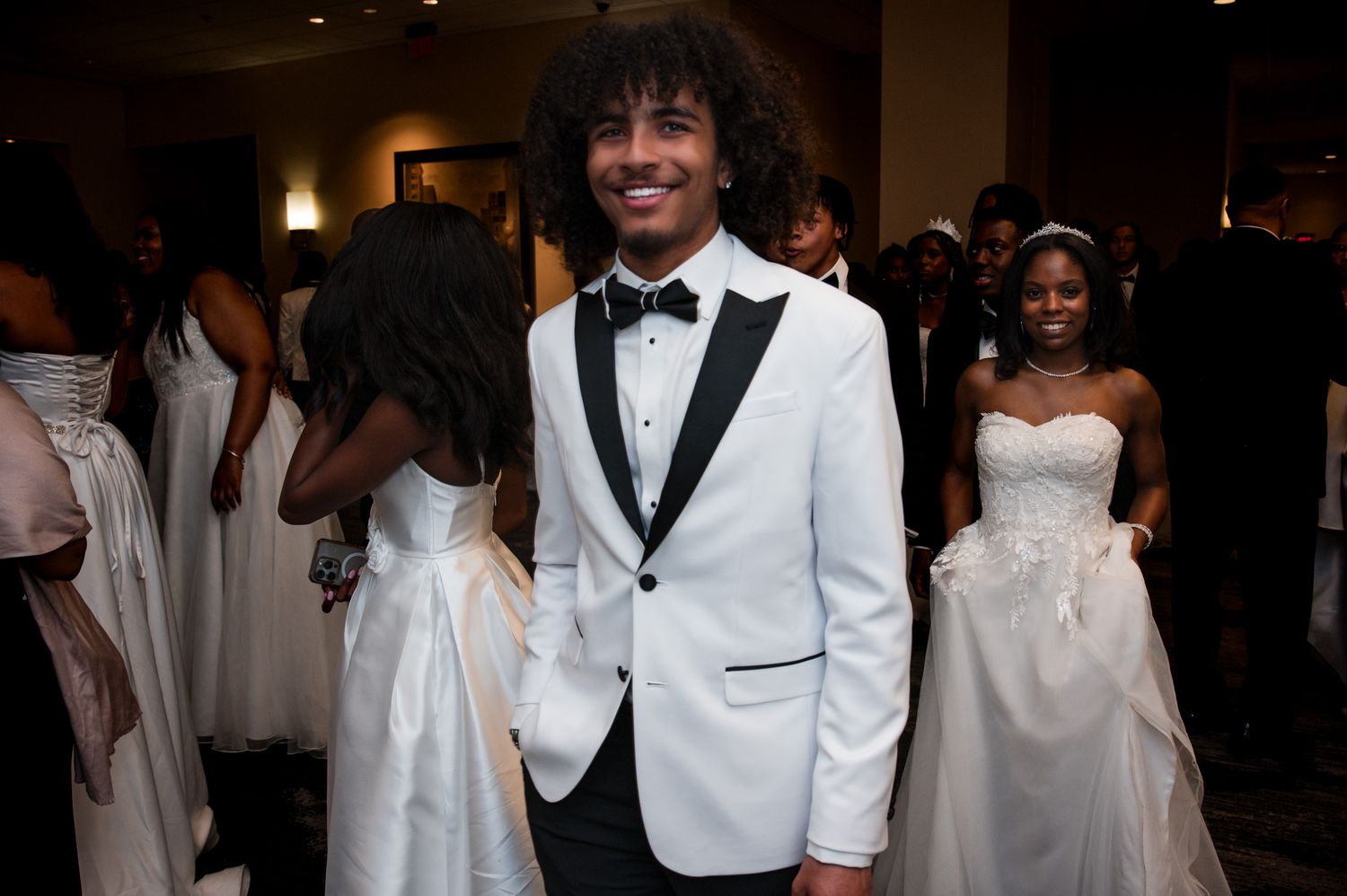 Young Black man in white tuxedo jacket and bow tie smiling at a formal debutante ball event.