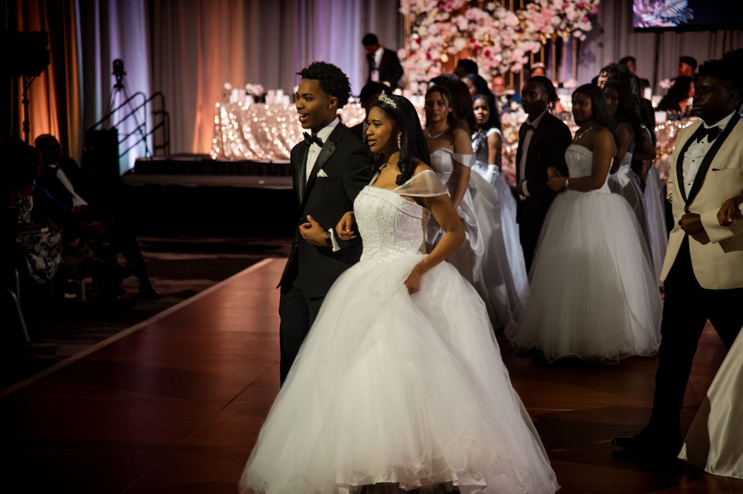 Young Black couple in formal attire dancing at an elegant debutante ball with floral stage backdrop.