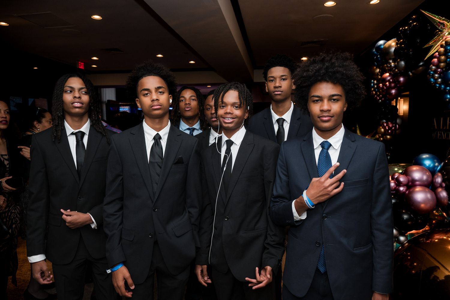 Group of young Black men in black suits posing together at a formal event with balloon decorations.