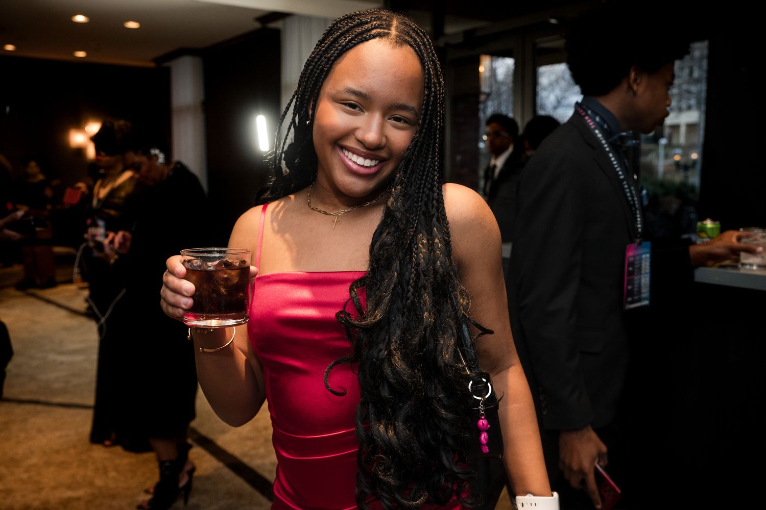 Young Black woman in red dress smiling while holding a drink at a formal social event.
