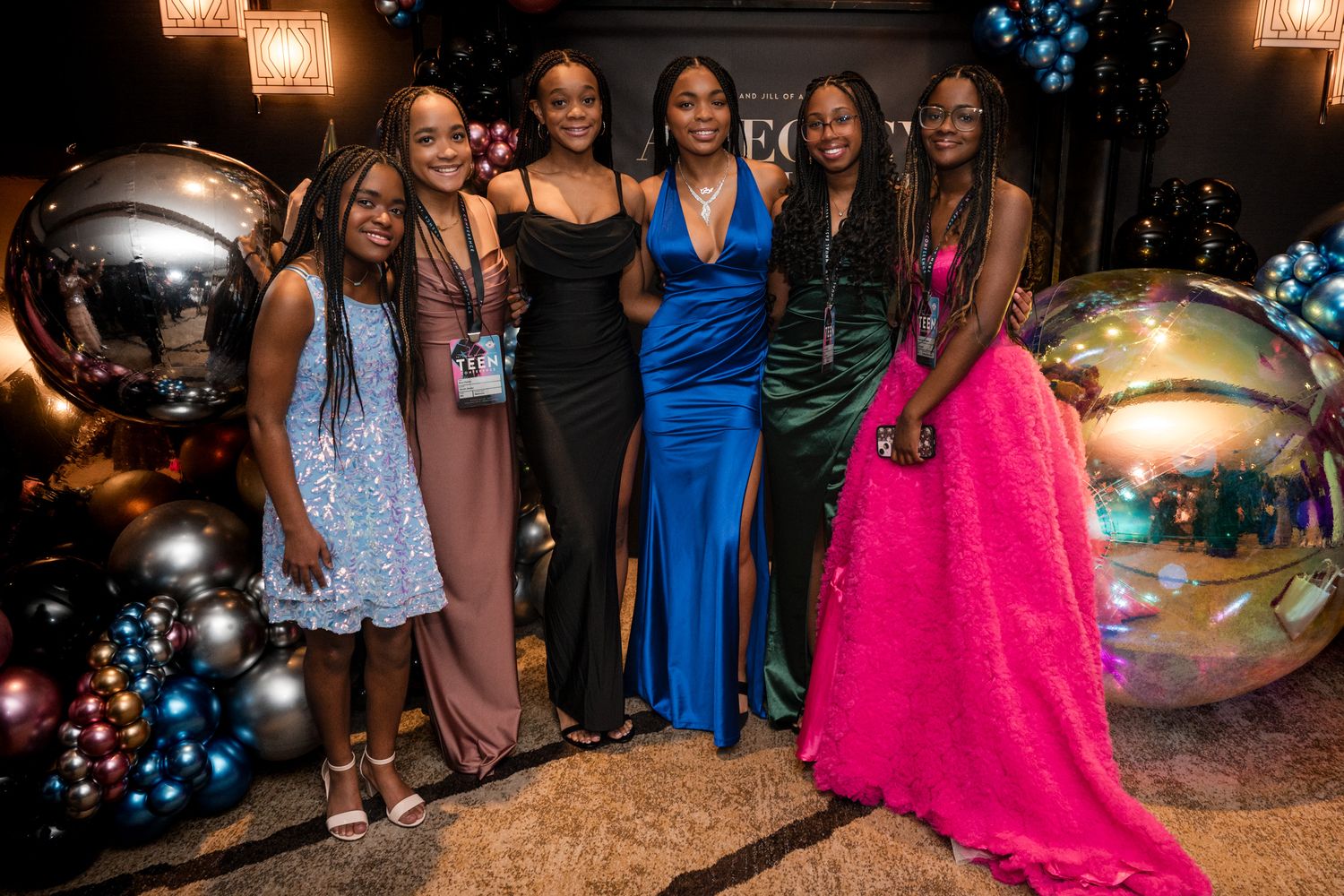 Group of six young Black women in colorful formal gowns posing together at an elegant social event.
