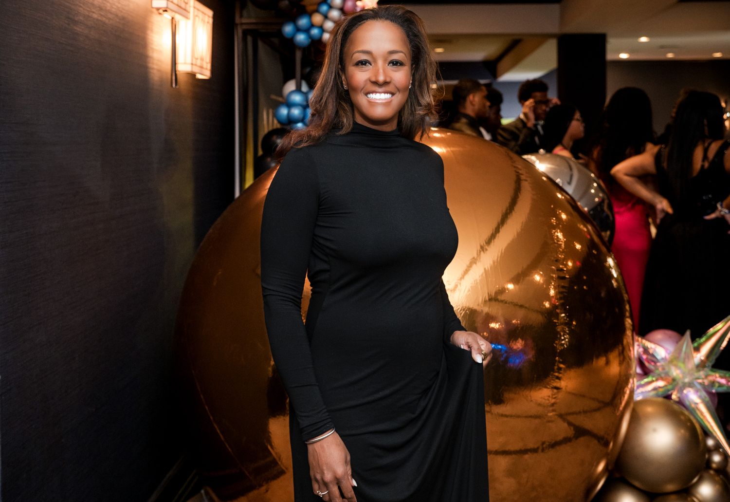 Woman in black turtleneck dress smiling at upscale event with gold balloon decor in background.