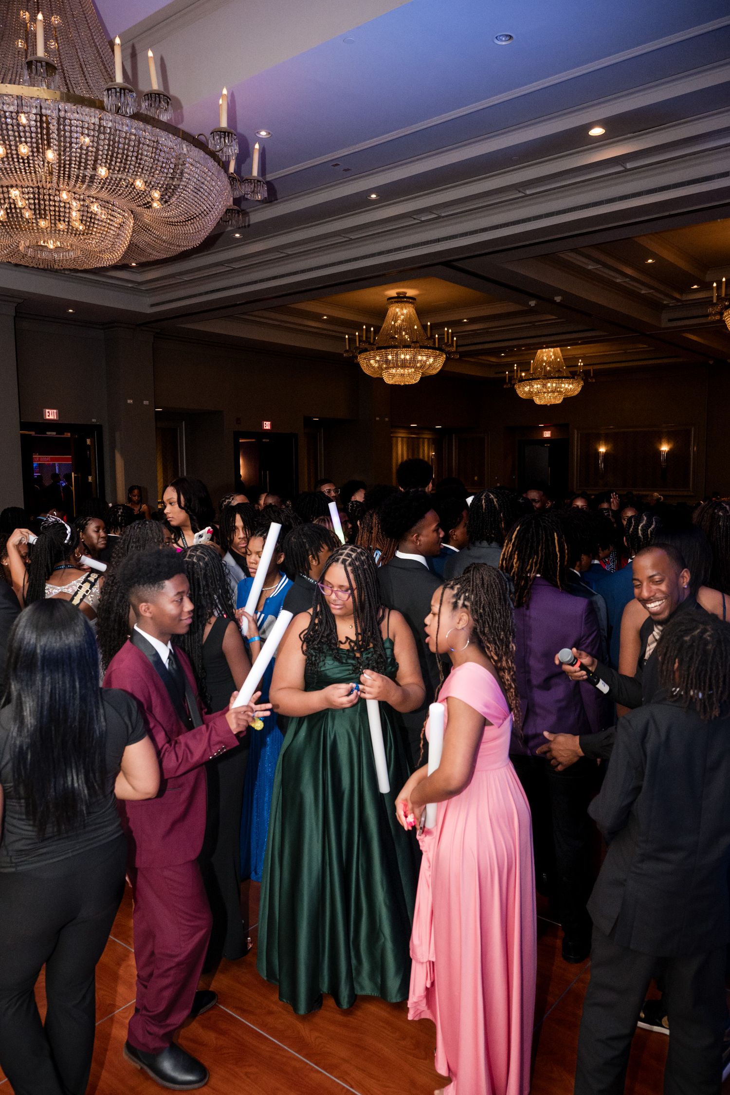 Teens in formal wear dancing under crystal chandeliers at an elegant prom or formal event.