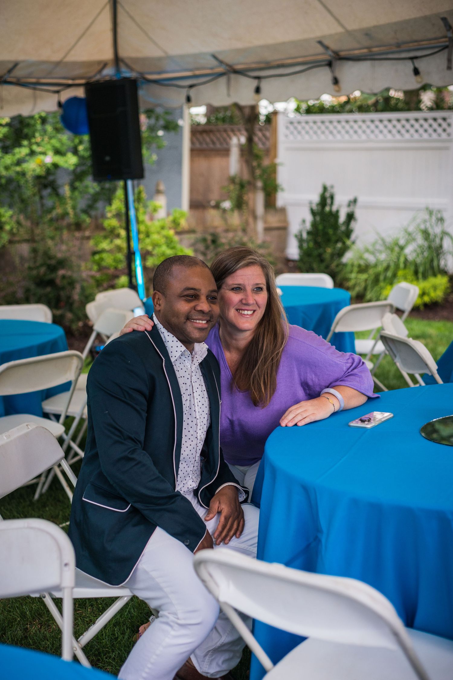 A smiling couple poses together at an outdoor event with blue tablecloths under a tent.