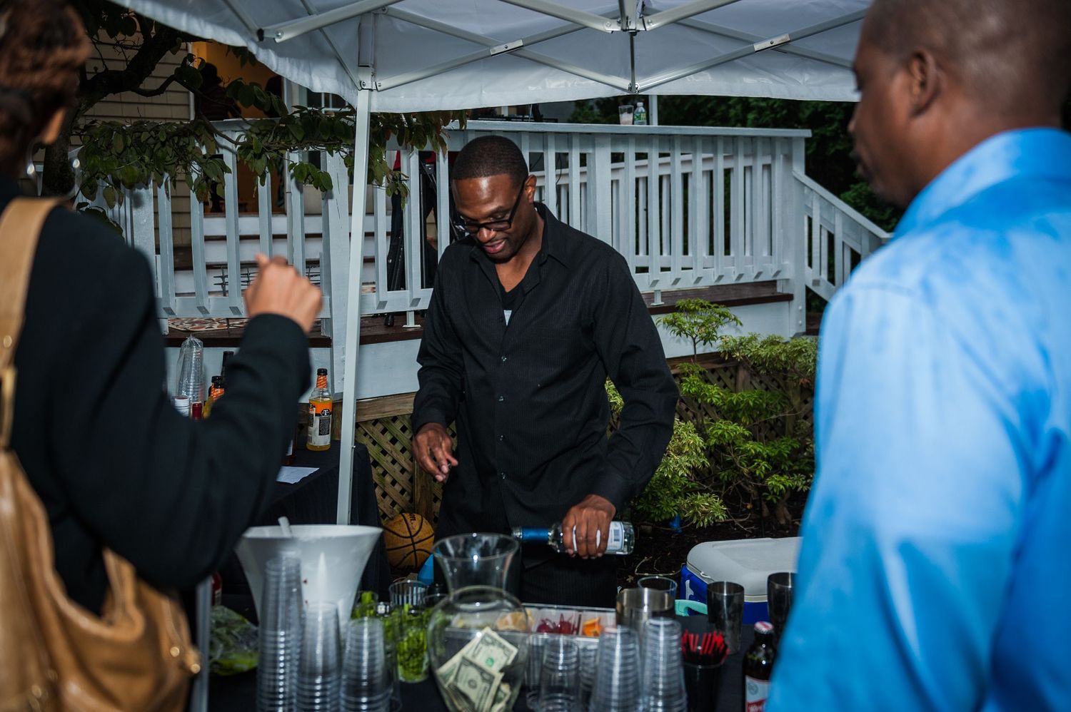 A bartender pours drinks for guests at an outdoor evening party under a tent.