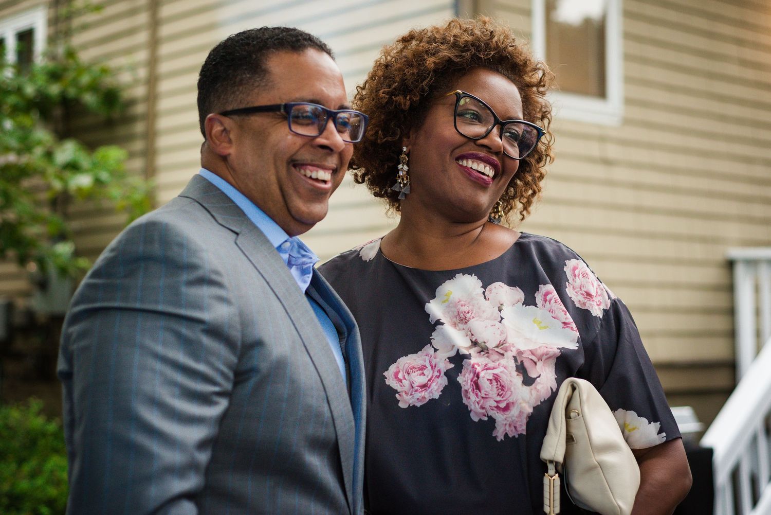 A well-dressed couple smiles and laughs together outside a house at a gathering.