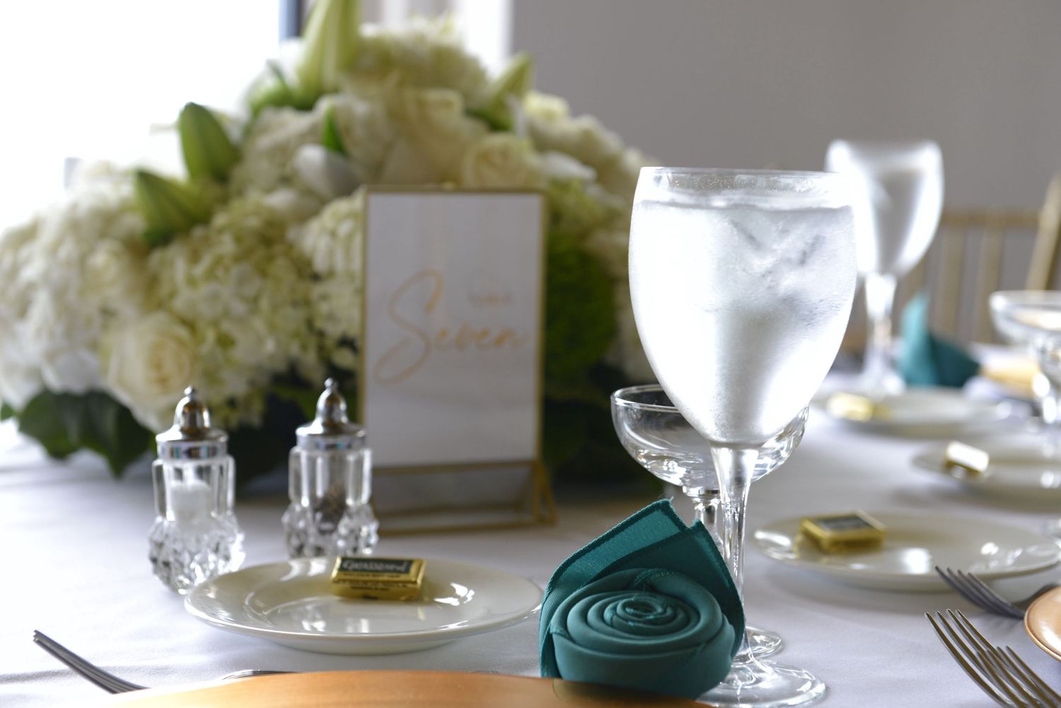 Elegant table setting with teal napkin, glassware, gold accents, and white floral centerpiece.