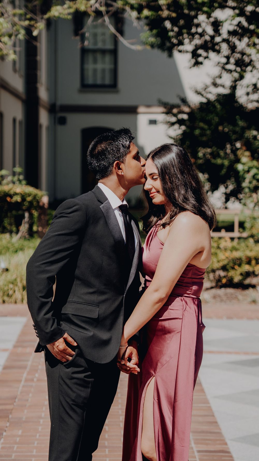 A romantic couple shares a tender moment outdoors, with the person in a dark suit leaning close to their partner in a burgundy gown.