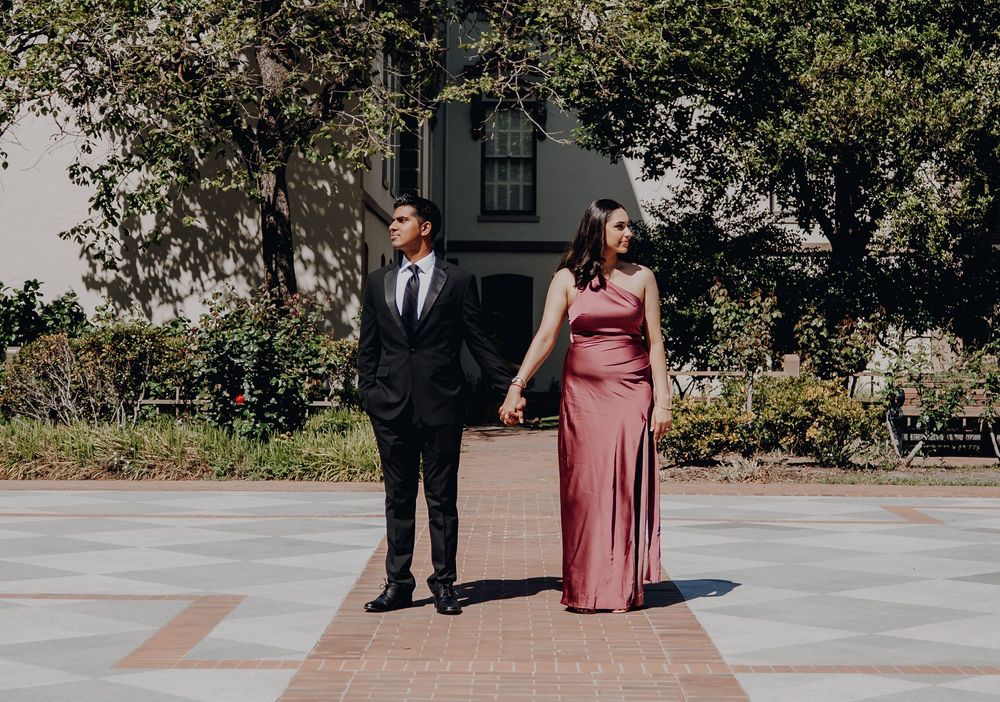 A couple stands together outdoors, with one wearing a dark suit and the other in a long pink formal dress.