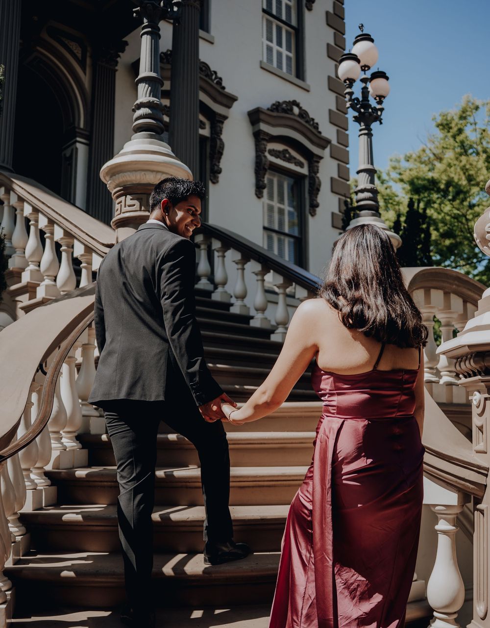 Couple in formal attire walking up elegant stone steps with ornate light posts and architectural details in sunlit setting.