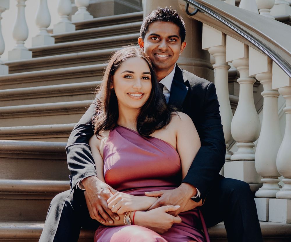 A couple poses together on an elegant staircase with white balusters, the woman wearing a burgundy one-shoulder dress.