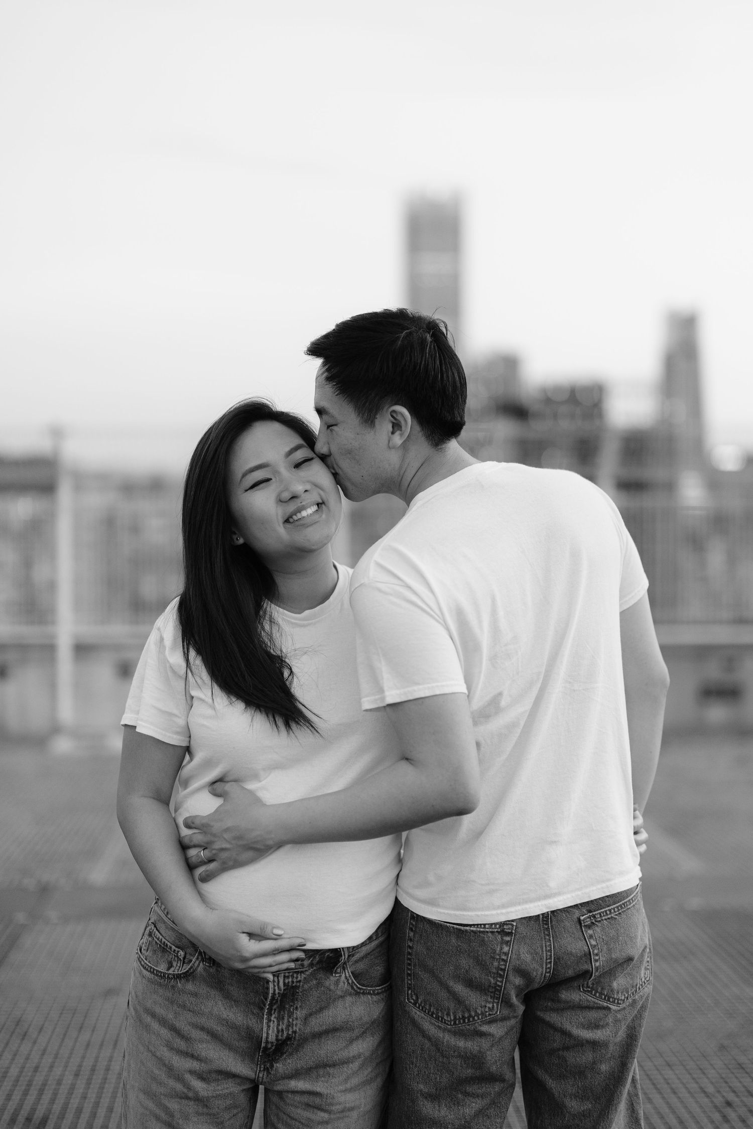A loving couple in white t-shirts and jeans embrace on a rooftop with a city skyline in the background, captured in black and white.