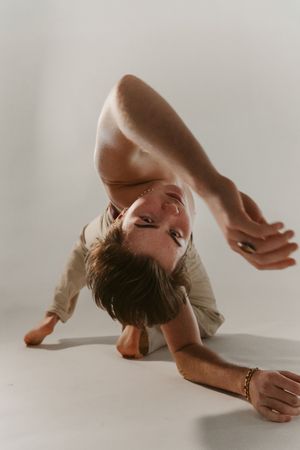 A flexible dancer bends gracefully on a white studio floor, wearing a beige outfit with a gold bracelet.